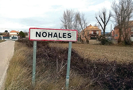 Cartel de entrada a la pedanía de Nohales junto a la carretera, con casas y árboles al fondo.